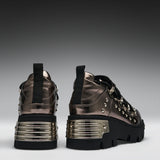 A pair of metallic bronze high-top platform sneakers with black soles, black laces, and silver studs, shown from the back and side against a dark gray gradient background.