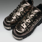 A pair of metallic bronze goth-style lace-up sneakers with silver studs, decorative metal hardware, and black laces, shown on a neutral gray background.