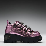 A metallic pink platform sneaker with black laces, silver studs, and a chunky black sole, shown from a side profile on a gray gradient background.
