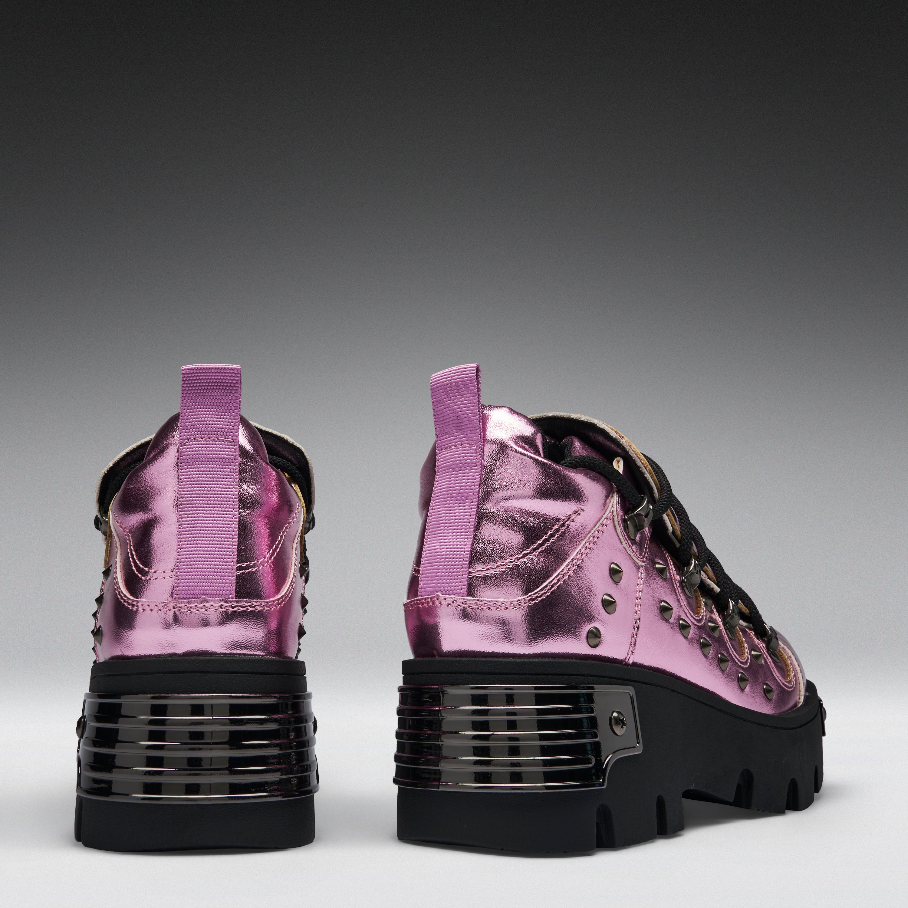 A pair of metallic pink high-top sneakers with chunky black platform soles, studded details, and purple pull tabs, shown from the back against a dark gray gradient background.