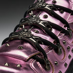 Close-up of a metallic pink high-top sneaker with black laces, silver conical studs, and ornate silver lace hardware featuring a wolf head design.