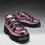 A pair of metallic pink platform sneakers with black lug soles, black decorative laces, and silver studs and hardware, photographed on a dark-to-light gray studio background.