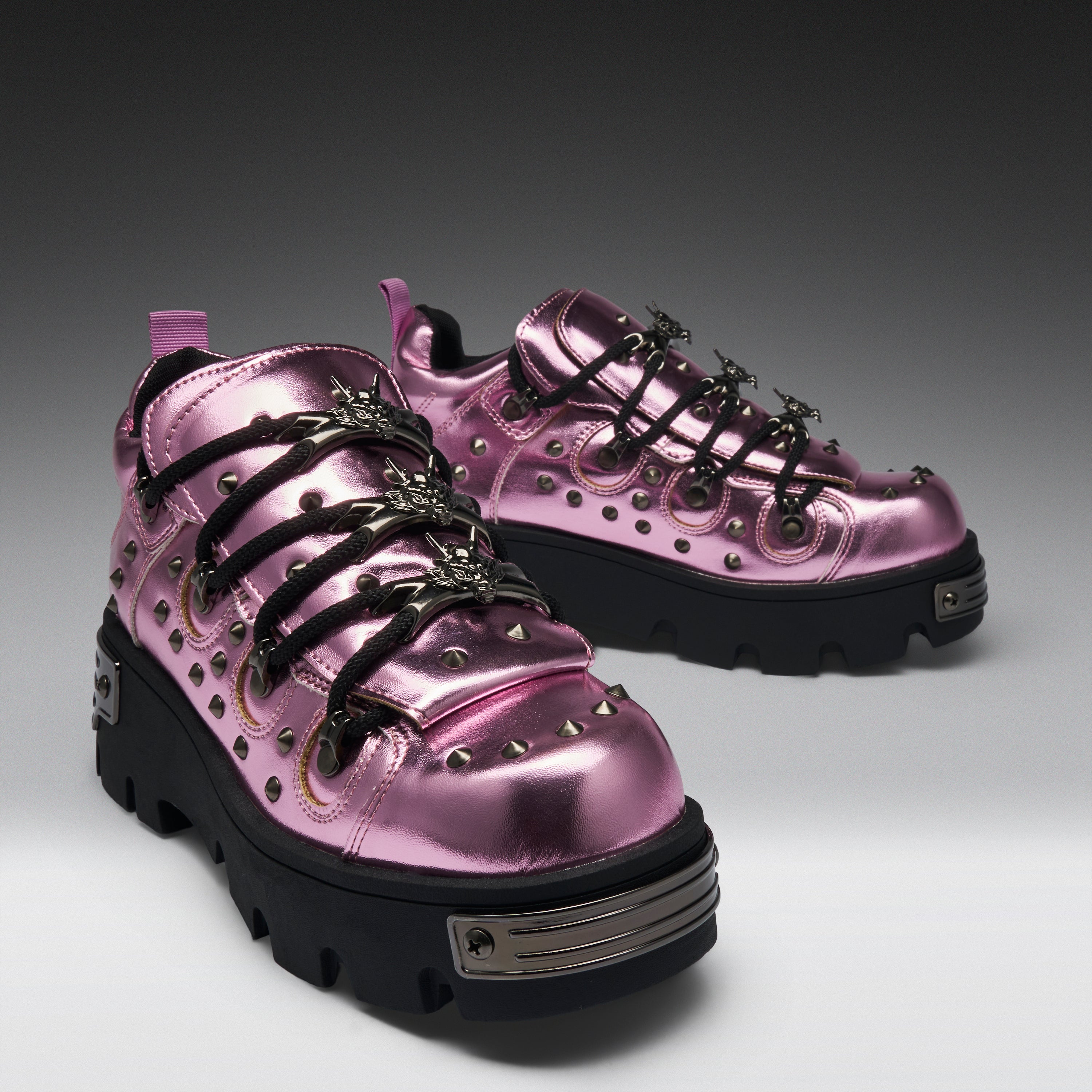 A pair of metallic pink platform sneakers with black lug soles, black decorative laces, and silver studs and hardware, photographed on a dark-to-light gray studio background.