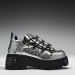 A metallic silver platform sneaker with black lug soles, black laces, and silver studs, shown from a side profile on a gray gradient background.