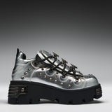 A metallic silver platform sneaker with black lug soles, black laces, and silver studs, shown from a side profile on a gray gradient background.