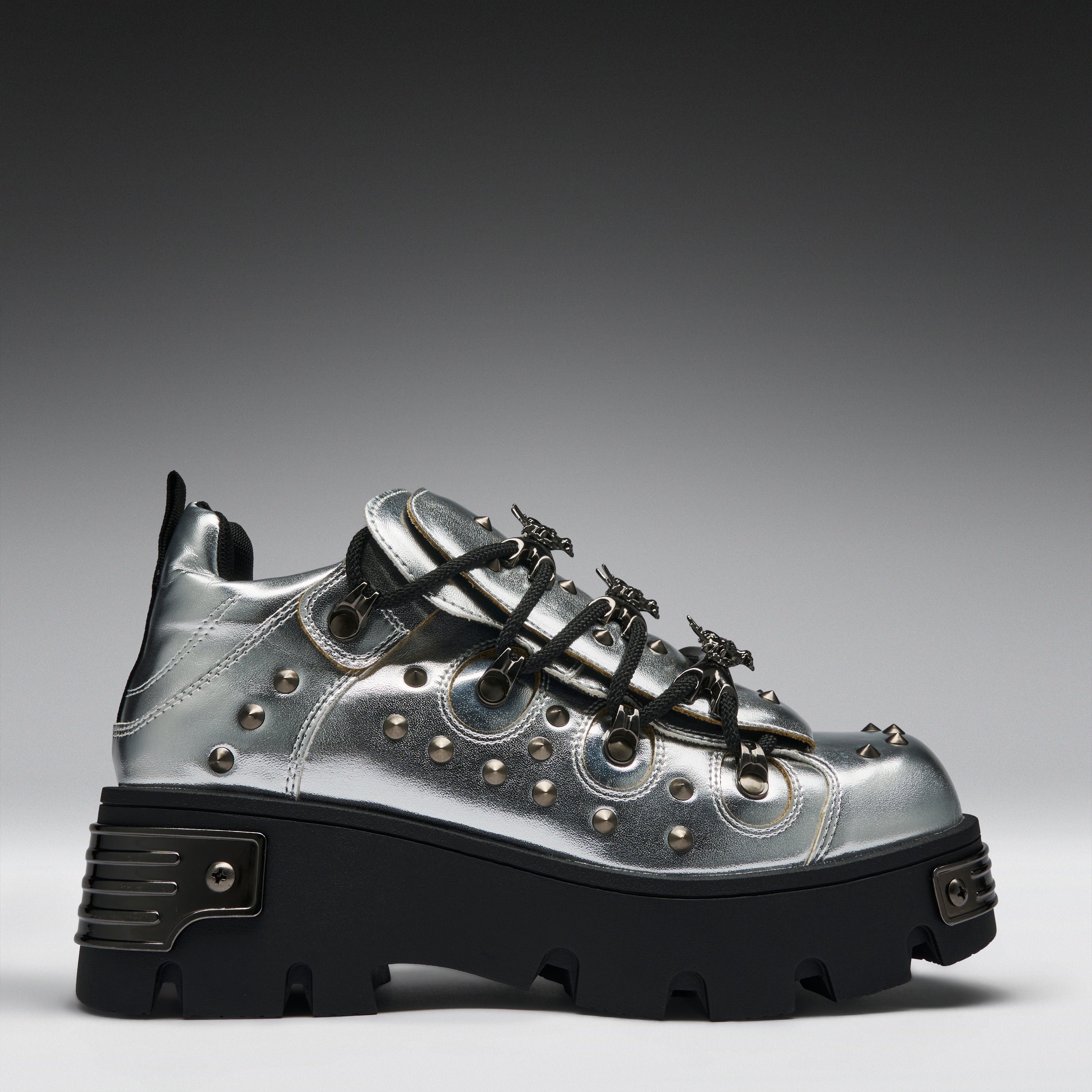 A metallic silver platform sneaker with black lug soles, black laces, and silver studs, shown from a side profile on a gray gradient background.