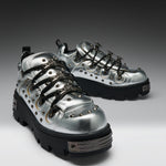 A pair of metallic silver high-top sneakers with black platform soles, black laces, and silver studs, displayed on a dark-to-light gray gradient studio background.