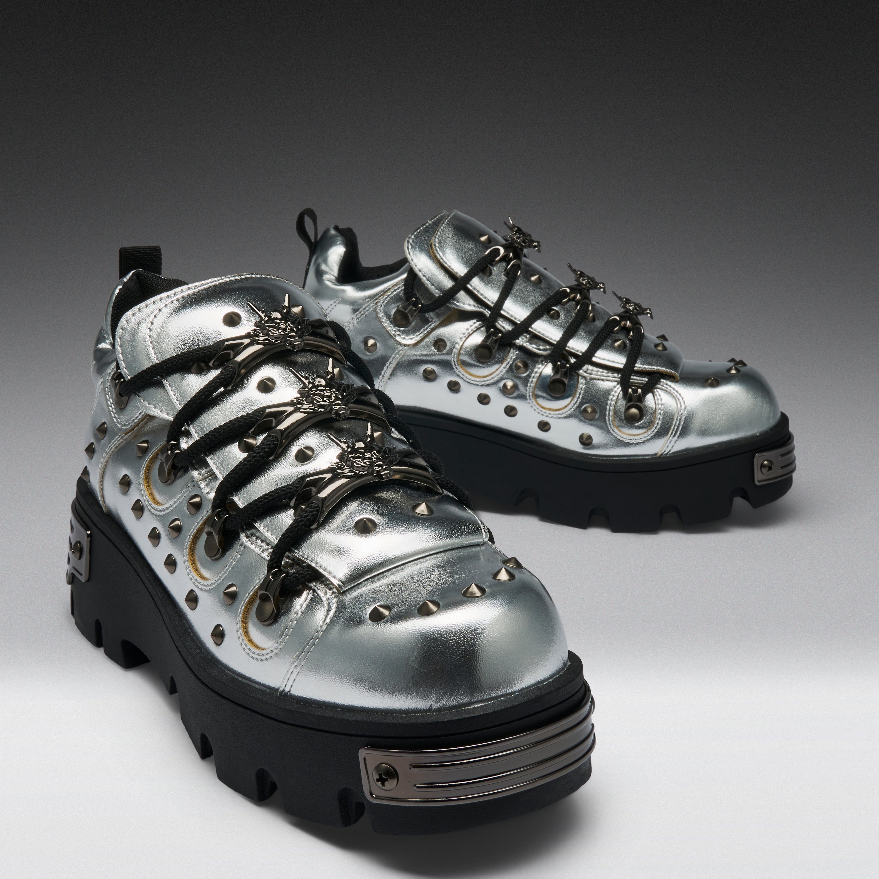 A pair of metallic silver high-top sneakers with black platform soles, black laces, and silver studs, displayed on a dark-to-light gray gradient studio background.