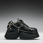 A black leather platform sneaker with chunky lug soles, silver chain details draped across the side, metal studs, and a small cross charm, shown against a dark gray gradient background.