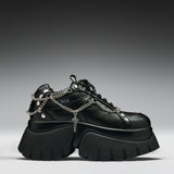 A black leather platform sneaker with chunky lug soles, silver chain details draped across the side, metal studs, and a small cross charm, shown against a dark gray gradient background.