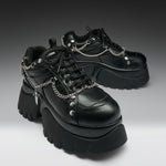 A pair of black leather platform sneakers with chunky lug soles, black laces, and silver chain details draped across the sides, photographed on a dark-to-light gray gradient studio background.