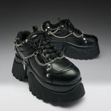 A pair of black leather platform sneakers with chunky lug soles, black laces, and silver chain details draped across the sides, photographed on a dark-to-light gray gradient studio background.