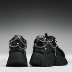 A pair of black chunky platform sneakers with silver chain and stud embellishments, shown from the back against a dark gray gradient background.
