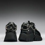 A pair of black chunky platform sneakers with silver chain and stud embellishments, shown from the back against a dark gray gradient background.