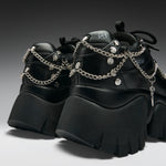 A pair of chunky black platform sneakers with silver chain straps, studs, and a small hanging cross charm, shown on a dark gray gradient studio background.