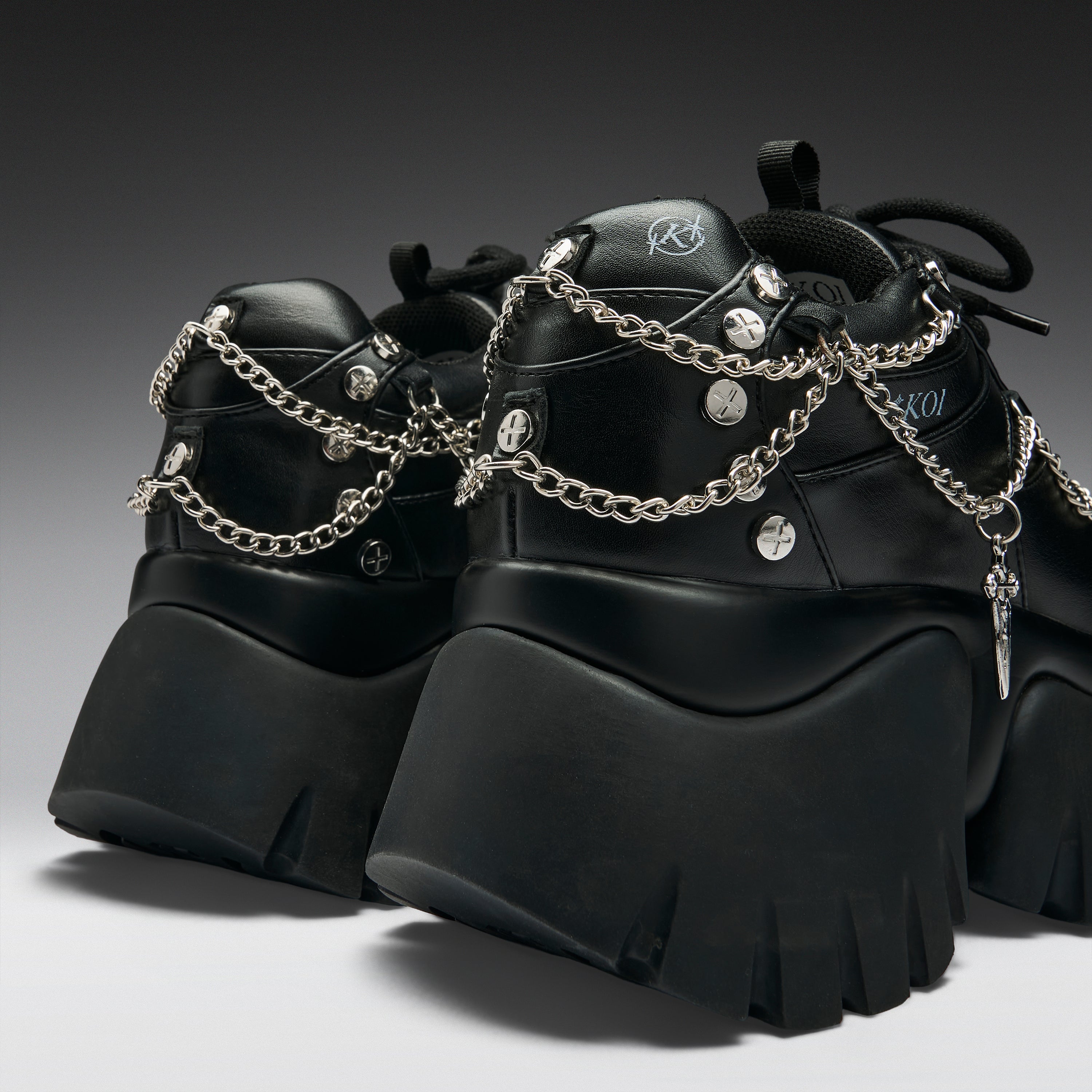 A pair of chunky black platform sneakers with silver chain straps, studs, and a small hanging cross charm, shown on a dark gray gradient studio background.