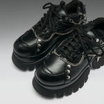 A pair of black leather platform sneakers with chunky lug soles, black laces, and silver chain details with skull and spike embellishments, shown on a gray gradient background.