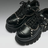 A pair of black leather platform sneakers with chunky lug soles, black laces, and silver chain details with skull and spike embellishments, shown on a gray gradient background.