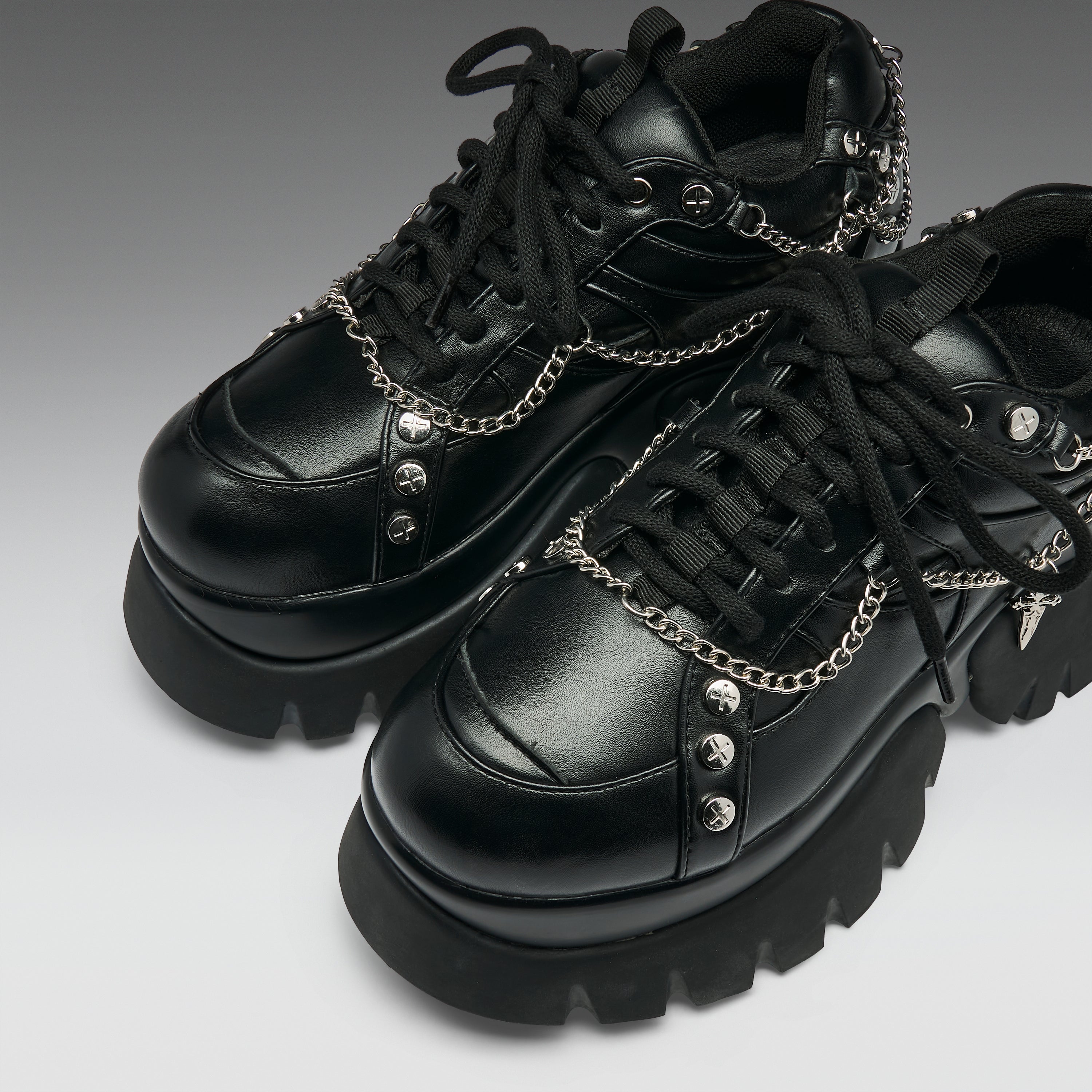 A pair of black leather platform sneakers with chunky lug soles, black laces, and silver chain details with skull and spike embellishments, shown on a gray gradient background.