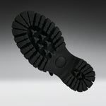 A close-up of a black rubber shoe sole, showing the deep lugs and tread pattern, photographed against a dark-to-light gray gradient background.