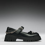 A black leather Mary Jane platform shoe with a chunky lug sole, a decorative tassel on the toe, and a silver chain draped from the buckle, shown against a dark gray gradient background.
