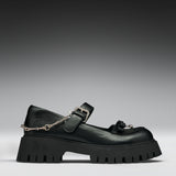 A black leather Mary Jane platform shoe with a chunky lug sole, a decorative tassel on the toe, and a silver chain draped from the buckle, shown against a dark gray gradient background.