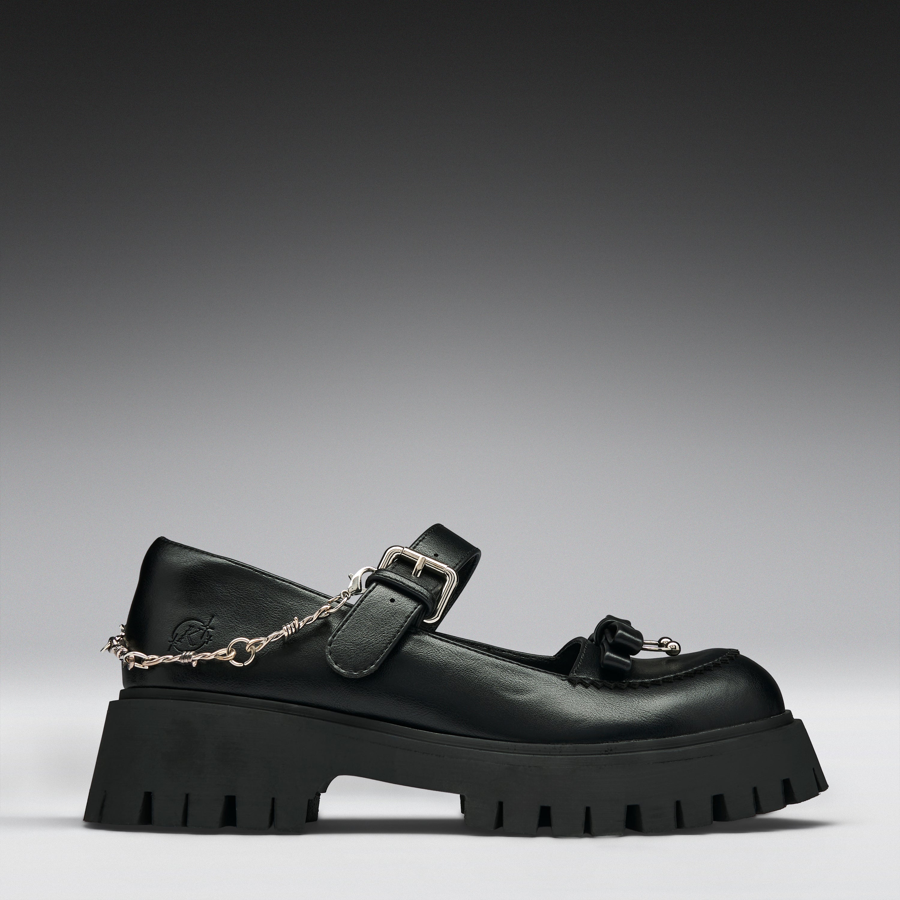 A black leather Mary Jane platform shoe with a chunky lug sole, a decorative tassel on the toe, and a silver chain draped from the buckle, shown against a dark gray gradient background.
