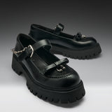 A pair of black leather Mary Jane platform shoes with chunky lug soles, decorative bow details, buckle straps, and silver-tone chain accents, photographed on a dark-to-light gray studio background.