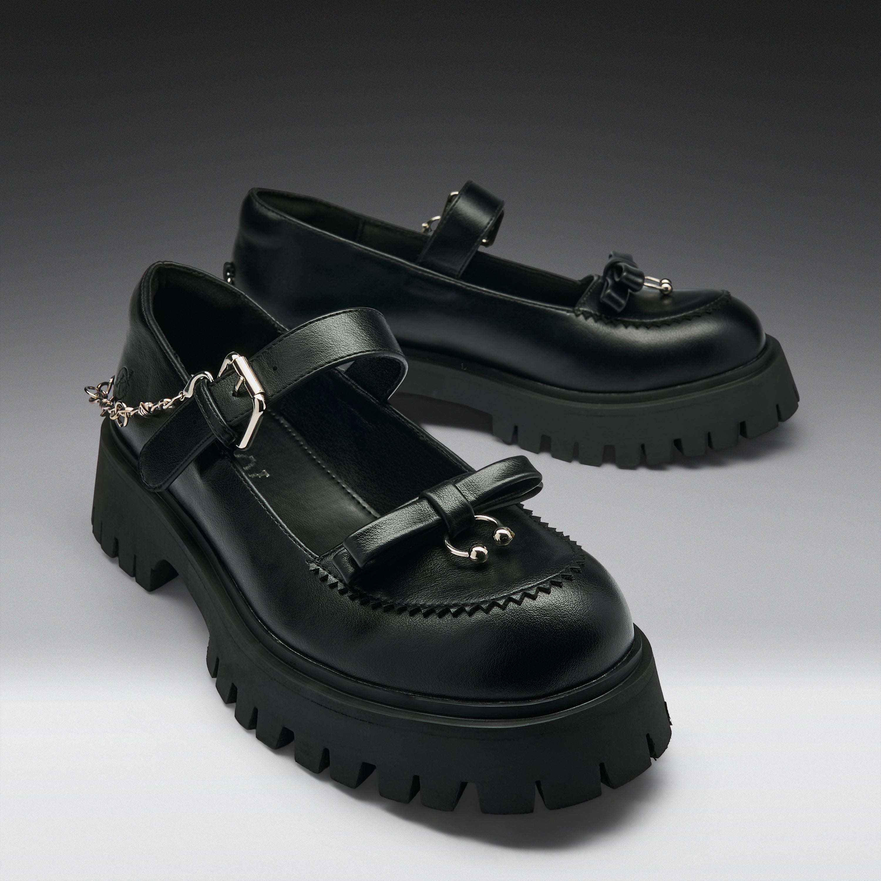 A pair of black leather Mary Jane platform shoes with chunky lug soles, decorative bow details, buckle straps, and silver-tone chain accents, photographed on a dark-to-light gray studio background.
