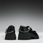 A pair of black leather Mary Jane shoes with chunky lug soles and silver chain details, shown from the back against a dark gray gradient background.