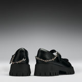 A pair of black leather Mary Jane shoes with chunky lug soles and silver chain details, shown from the back against a dark gray gradient background.