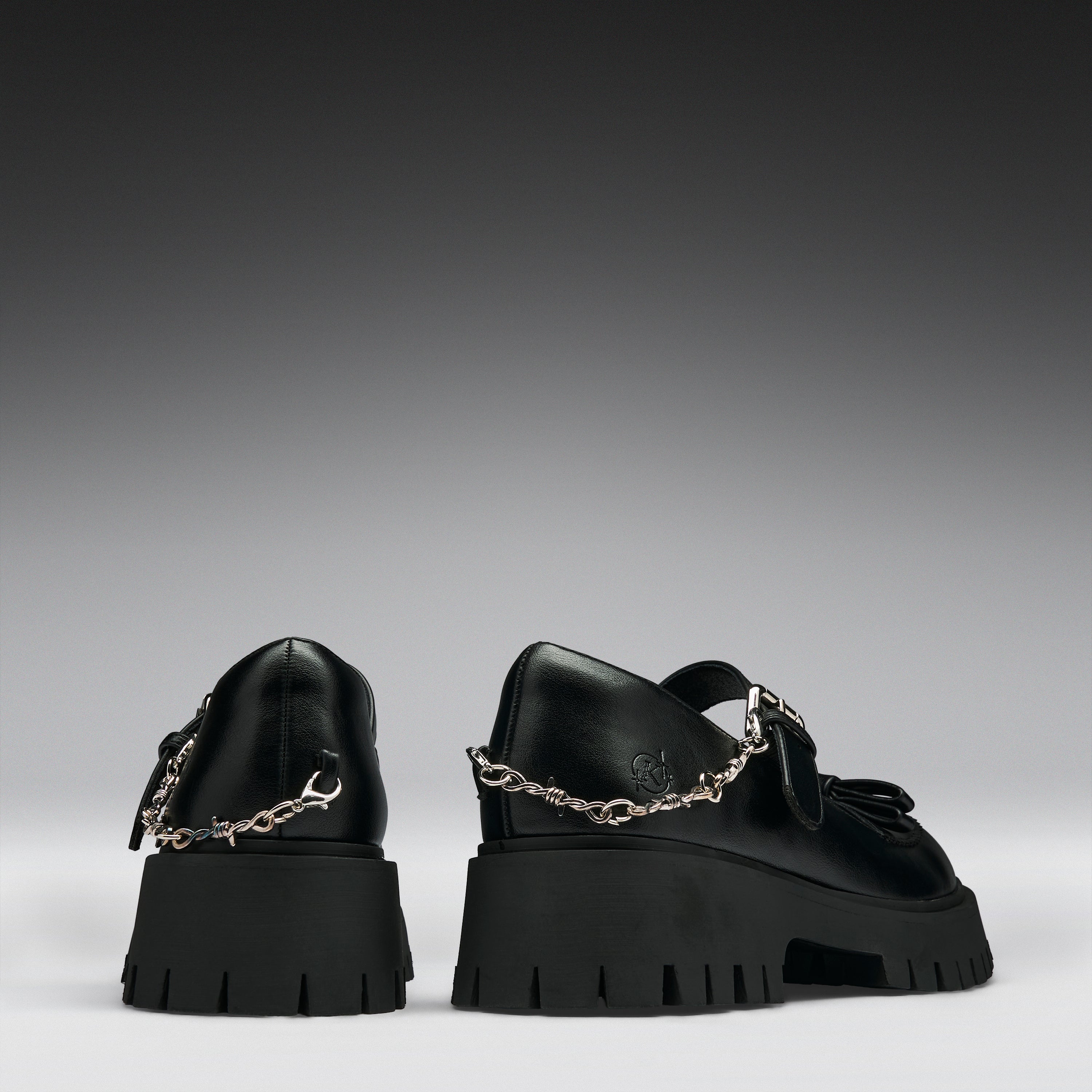 A pair of black leather Mary Jane shoes with chunky lug soles and silver chain details, shown from the back against a dark gray gradient background.