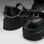 Black leather platform mule shoes with chunky lug soles, decorative stitching, a bow detail, and a silver chain accent on the strap, shown on a clean light gray studio background.