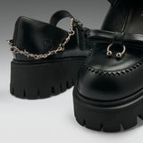 Black leather platform mule shoes with chunky lug soles, decorative stitching, a bow detail, and a silver chain accent on the strap, shown on a clean light gray studio background.