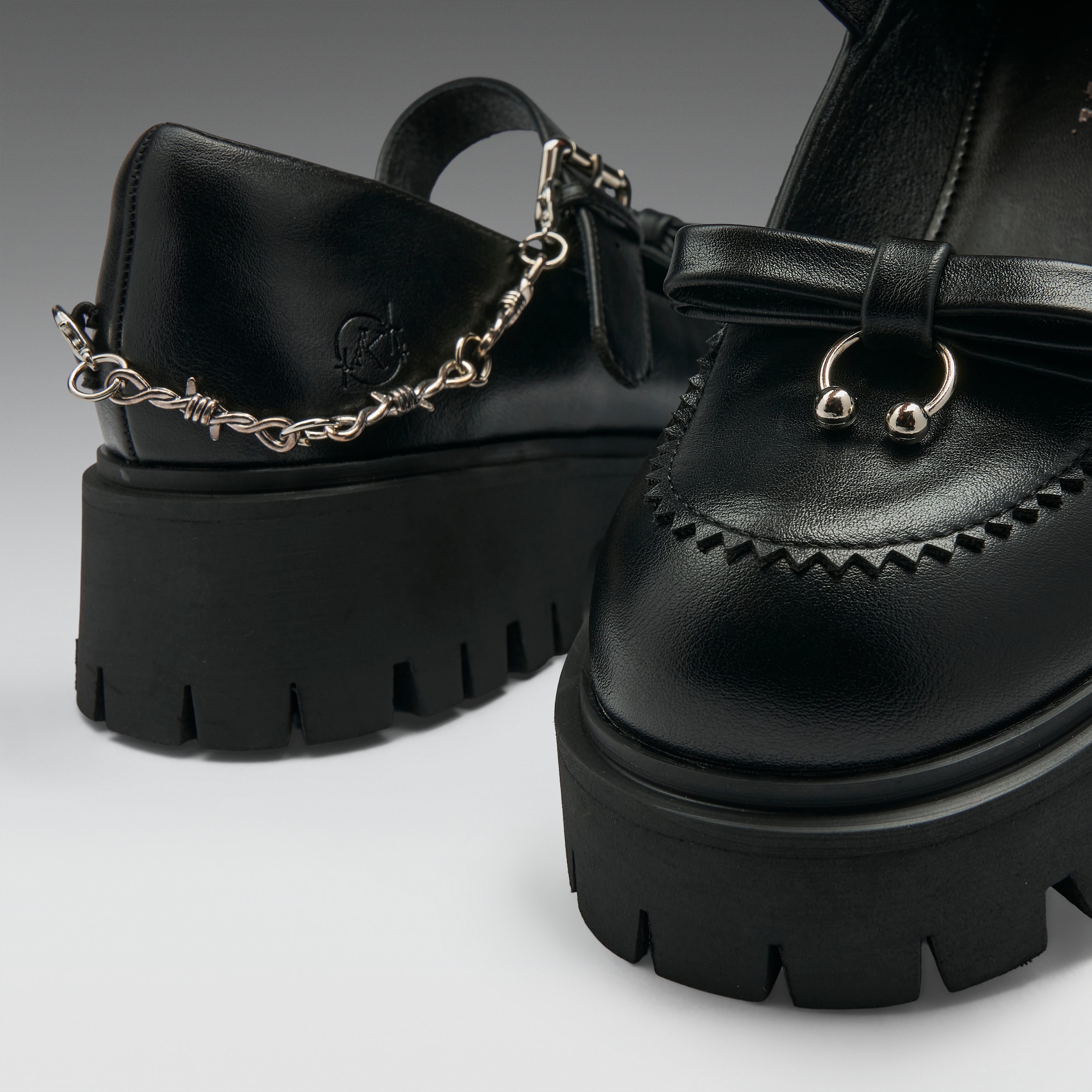 Black leather platform mule shoes with chunky lug soles, decorative stitching, a bow detail, and a silver chain accent on the strap, shown on a clean light gray studio background.