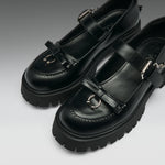 A pair of black leather Mary Jane-style shoes with chunky lug soles, decorative bow details, and silver-tone metal hardware, photographed on a clean gray studio background.