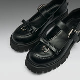 A pair of black leather Mary Jane-style shoes with chunky lug soles, decorative bow details, and silver-tone metal hardware, photographed on a clean gray studio background.